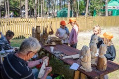 Dzieci stoją przy stole, na którym prezentowane są rzeźby. Z drugiej strony stołu siedzi mężczyzna i kobieta, którzy wykonują kolejne rzeźby. W tle drewniany płot, drzewa, namioty twórców i drewniana zabudowa w Zagrodzie Kurpiowskiej..