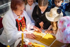 Dzieci przyglądają się pracy kobiety w stroju kurpiowskim wykonującej świece z wosku pszczelego