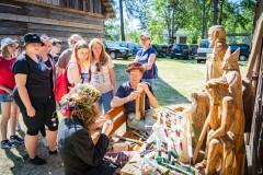 Grupa widzów wokół rzeźbiarza siedzącego przy stole, na którym ustawione są rzeźby. W tle fragment drewnianego budynku oraz zielony teren, zaparkowane samochody, drzewa.