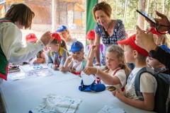 Dzieci siedzą przy przy stole w namiocie. Przy dzieciach stoi opiekunka. Po drugiej stronie stołu stoi kobieta ucząca haftowania.