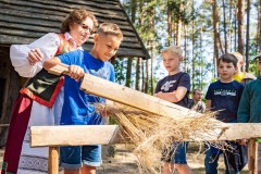 Chłopcy w asyście kobiety w stroju kurpiowskim uczą się obróbki lnu