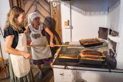 Dwie kobiety stoją przy zabytkowym piecu w chacie kurpiowskiej. Jedna z kobiet przy pomocy specjalnej łopaty wyjmuje z pieca blachy z upieczonym „łogodniakiem” i układa je na kuchni.