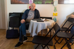 Mężczyzna siedzący przy okrągłym stoliku. Za nim ściana, na której widać fragmenty fotografii oprawionych w ramy