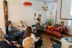 Widzowie siedzący na sali patrzą w stronę pracownicy Muzeum prezentującej jedną z pamiątek z podróży do Afryki. Za nią stolik z pamiątkami, dalej rollup z logotypem MKK oraz Marki Mazowsze. Pod sufitem wiszą kierce.