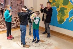 Dzieci przymierzają kolorowe maski wykonane z papieru. W tle fragment fotografii afrykańskiej wioski  oraz mapy Afryki.