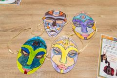 Cztery kolorowe maski z papieru oraz fragmenty kart edukacyjnych leżące na stole.