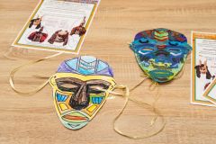 Dwie kolorowe maski z papieru oraz fragmenty kart edukacyjnych leżące na stole.