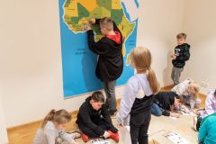 Niektóre dzieci siedzą na ziemi rozwiązując karty edukacyjne, inne stoją. Chłopiec przymocowuje papierowe postacie do dużej mapy Afryki wiszącej na ścianie.