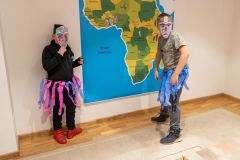 Dwóch chłopców w maskach z papieru i stroju z pasków bibuły pozuje na tle dużej mapy Afryki.