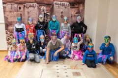 Grupa dzieci w papierowych maskach i strojach z kolorowych pasków bibuły pozuje do zdjęcia na tle dużej fotografii przedstawiającej wioskę Dogonów.