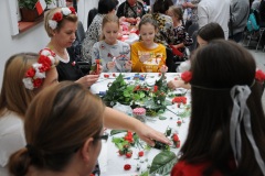 Dziewczynki wraz z Kobietami wykonują wianki z biało-czerwonych kwiatów.