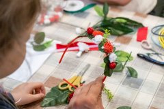 Dziewczynka siedzi przy stole, siedząca obok niej pani  trzyma w lewej ręce opaskę zrobioną z biało czerwonych różyczek i zielonych listków, w tle leżą materiały potrzebne do zrobienia opaski     