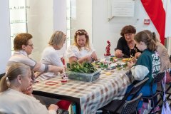 Dwie pracownice muzeum gdzie jedna ma opaskę na głowie z biało czerwonych różyczek, pokazują pozostałym jak zrobić opaski, cztery kobiety w różnym wieku i dwie nastolatki siedzą przy stole gdzie nastolatka trzyma w ręce nożyczki wycinając listki potrzebne do zrobienia  opaski, kobieta obok trzyma gotową opaskę z biało czerwonych różyczek. W tle widać flagę biało czerwoną.