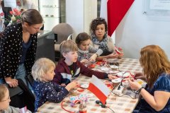 Siedząca przy stole pracownica muzeum pokazuje czwórce dzieci, jednej kobiecie siedzącej, jednaj stojącej jak wykonać i przykleić ozdobne wstążeczki na słoiczek w barwach biało czerwonych. Na stole leżą wstążki różnej szerokości, nożyczki, małe pistolety do klejenia na gorąco. Temu wszystkiemu towarzyszy stojąca w słoiczku flaga Polski, w tle również wisi duża flaga o barwach narodowych. 