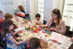 Przy dużym stole siedzą dwie kobiety, dwie dziewczynki i trzech chłopców, przy jednym z chłopców stoi sklejony lampion przedstawiający budynek Muzeum Kultury Kurpiowskiej, pozostałe dzieci zaczynają klejenie, na pierwszej połowie stołu leżą materiały potrzebne do klejenia, taśma dwustronna, klej, nożyczki, oraz taśma biało czerwona do zrobienia flagi Polski. Na środku stołu stoi w mały flakonik z małą flaga która została zrobiona przez uczestników warsztatów. Na końcu stołu siedząca kobieta klei za pomocą pistoletu na gorąco. W tle widać trzy gabloty z figurkami żołnierzyków.
