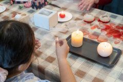 Siedząca przy stole dziewczynka trzyma w ręce biały tiul z którego będzie wykonany kokarda biało czerwony, obok leżą wycięte kawałki tiulu, stoją dwie płonące świeczki.  