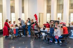 Grupa osób biorących udział w warsztatach muzealnych wykonuje ozdoby i kokardy w barwach naszego państwa, w tle widoczna duża flaga Polski i  cztery małe stojące na stołach. Przy pierwszym stole stoi pani Dyrektor muzeum a w tle widoczna stojąca przy komputerze  pracownica muzeum.