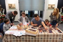 Po prawej stronie stołu stojąca kobieta, mały chłopczyk i siedzący mężczyzna układają puzzle. Obok nich  po lewej stronie stoi dziewczynka z opaska na głowie w barwach  naszego państwa obok stoi pochylony mężczyzna i siedzący chłopczyk przy stole gdzie leżą kartki papieru do kolorowania i kredki w pudełku. W tle po lewej i prawej stronie stoją kwiaty biało czerwone i wiszą obrazy na ścianie.