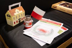 Na brzegu stołu leżą dwie kartki papieru do kolorowania za nimi stoi słoiczek obklejony barwami narodowymi a w nim włożona na patyczku  mała flaga Polski  za nią stoi lampion przedstawiający budynek Muzeum Kultury Kurpiowskiej, w tle leżą puzzle w pudełku.