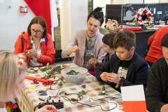 Dwie kobiety jedna dziewczynka i chłopiec siedzą przy stole i robią opaski na głowę z białych różyczek, zielonych listków i czerwonych jagód żurawiny. Na stole stoi koszyczek z listkami, leży czerwona bibuła karbowana, pistolet do klejenia na gorąco, nożyczki i mała flaga biało czerwona Polski. Za nimi wisi duża flaga Polski, a po prawej stronie w tle widoczne są lampiony i stroik z biało czerwonych kwiatów.