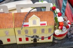 Zdjęcie przedstawia dwa lampiony z papieru przedstawiające budynek Muzeum kultury Kurpiowskiej, udekorowany słoiczek różyczkami w barwach biało czerwonych i opaskę na głowę z kwiatków w kolorze białym i czerwonym oraz zielonych listków. 