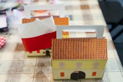 Na stole stoją dwa lampiony z papieru przedstawiające budynek Muzeum Kultury Kurpiowskiej a po między nimi włożona jest mała flaga biało czerwona Polski.