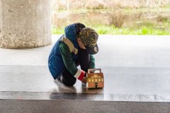 Przed budynkiem mauzoleum na schodach chłopczyk w czapce i niebieskiej kurtce postawił lampion klęcząc na jedno kolano. W tle widać betonowy słup i zielono szarą trawę.