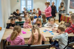 Dzieci siedząc przy stole wykonują  tradycyjne ozdoby choinkowe. Z prawej strony stoją dwie pracownice Muzeum i opiekunka grupy. Jedna opiekunka grupy siedzi z dziećmi przy stole.