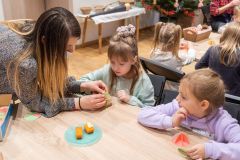 Dziewczynki siedząc przy stole wykonują tradycyjne ozdoby choinkowe z opłatka, tzw. światy.  Z lewej strony pracownica Muzeum pomaga jednej z dziewczynek.
