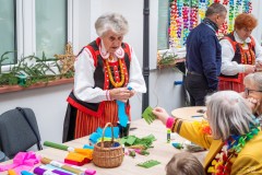 Dwie twórczynie kurpiowskie stoją przy swoich stołach, przy których siedzą uczestnicy warsztatów. W oddali przy jednej z twórczyń stoi mężczyzna 