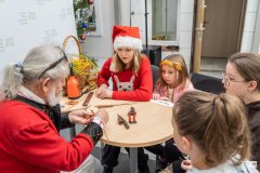 Cztery dziewczynki,  jedna dziewczynka w czapce Mikołaja, siedząc przy okrągłym stoliku przyglądają się pracy twórcy wykonującego ozdobę z wikliny