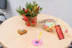 Na okrągłym stoliku stoi stroik świąteczny, gwiazdka z bibuły – zawieszka na choinkę, świat z opłatka zapakowany w folię z bilecikiem oraz niewielki pierniczek w kształcie serca