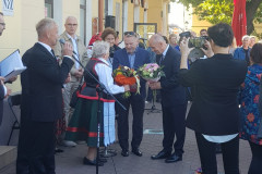 Ponad dwadzieścia osób stoi przed budynkiem. Mężczyzna wręcz kwiaty kobiecie w stroju kurpiowskim