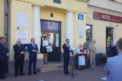 Kilkanaście osób stoi przed budynkiem. Na schodkach przed wejściem stoi kobieta z bębnem. Jeden z mężczyzn stoi przy mikrofonie, niedaleko kobieta w stroju kurpiowskim z bukietem kwiatów.