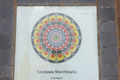 Na ziemi, zamontowana w chodniku plansza z kolorową, okrągłą wycinanką w stylu łowickim, poniżej wycinanki napis „Czesława Marchewka w kategorii twórczość ludowa”. W prawym górnym rogu planszy niewielki napis „ROK 2021”