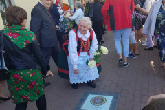 Przed budynkiem stoi kilkanaście osób. Kobieta w stroju kurpiowskim, trzymająca w dłoni jasnożółte róże pochyla się nad wmontowaną w chodnik planszą z kolorową wycinanką.