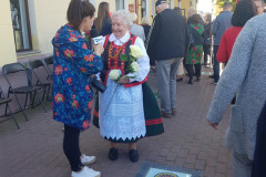Przed budynkiem kobieta w stroju kurpiowskim, trzymająca w dłoni jasnożółte róże udziela wywiadu kobiecie z mikrofonem. W tle kilkanaście innych osób