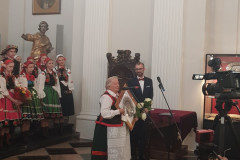 W stylowej sali kobieta w stroju kurpiowskim przemawia do mikrofonu, dalej stoi mężczyzna w garniturze. Z lewej strony chór składający się z osób w strojach ludowych, z prawej kamery