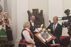W stylowej sali mężczyzna w garniturze wręcza kobiecie w stroju kurpiowskim okrągłą wycinankę oprawioną w ramę. Obok inny mężczyzna w garniturze. Z prawej strony fragment kamery. Z lewej strony dwie kobiety w strojach ludowych.