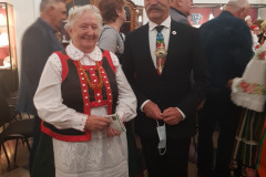 Kobieta w stroju kurpiowskim pozuje do zdjęcia z mężczyzną w garniturze. W tle kilka innych osób.