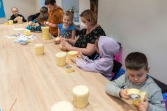 Fragment stołu przy którym siedzą dzieci. Wykonują bębenki  z plastikowych pojemników i papierowej taśmy. Kobieta w maseczce pomaga jednej z dziewczynek.