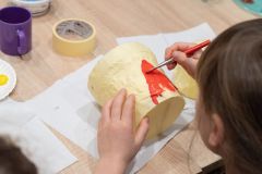 Dziecko siedząc przy stole wykonuje bębenek z plastikowych pojemników i papierowej taśmy.