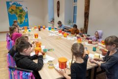 Wokół dużego stołu siedzi wiele dzieci. Wykonują bębenki  z plastikowych pojemników i papierowej taśmy. W tle na ścianie wisi duża mapa Afryki.