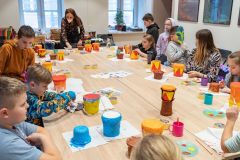 Wokół dużego stołu siedzi wiele dzieci. Wykonują bębenki  z plastikowych pojemników i papierowej taśmy. Pomagają im dwie kobiety w maseczkach.