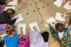 Dzieci siedzą na ziemi tworząc okrąg, rozwiązują karty edukacyjne. Widok fragmentu z góry.
