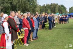 Uczestnicy uroczystości stoją obok miejsc siedzących w plenerze