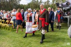 Uczestnicy uroczystości stoją obok miejsc siedzących w plenerze. Kobieta i mężczyzna w strojach kurpiowskich niosą duży chleb.