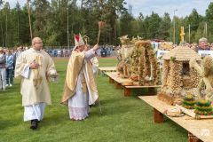 Biskup święci wieńce dożynkowe w plenerze. Obok ksiądz niesie naczynie z wodą święconą. W tle inni ludzie, uczestnicy uroczystości