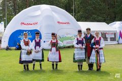 Pięć kobiet i mężczyzna w strojach kurpiowskich pozują do zdjęcia. W tle namioty z logiem Marki Mazowsze