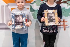 Chłopiec i dziewczynka trzymając w dłoniach ramkę ze zdjęciem pozują na tle dużej ilustracji przestawiającej dworek. Chłopiec ma na głowie czarny, papierowy cylinder